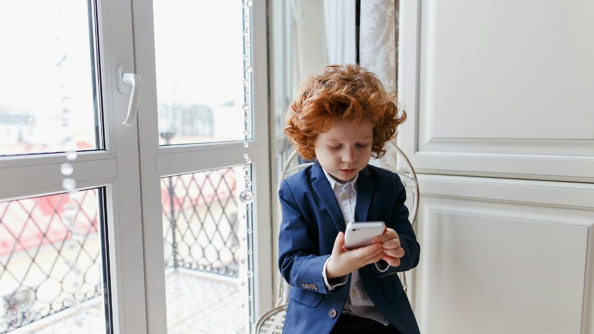 Red-haired boy wearing a suit using a smartphone indoors, seated near a window.
