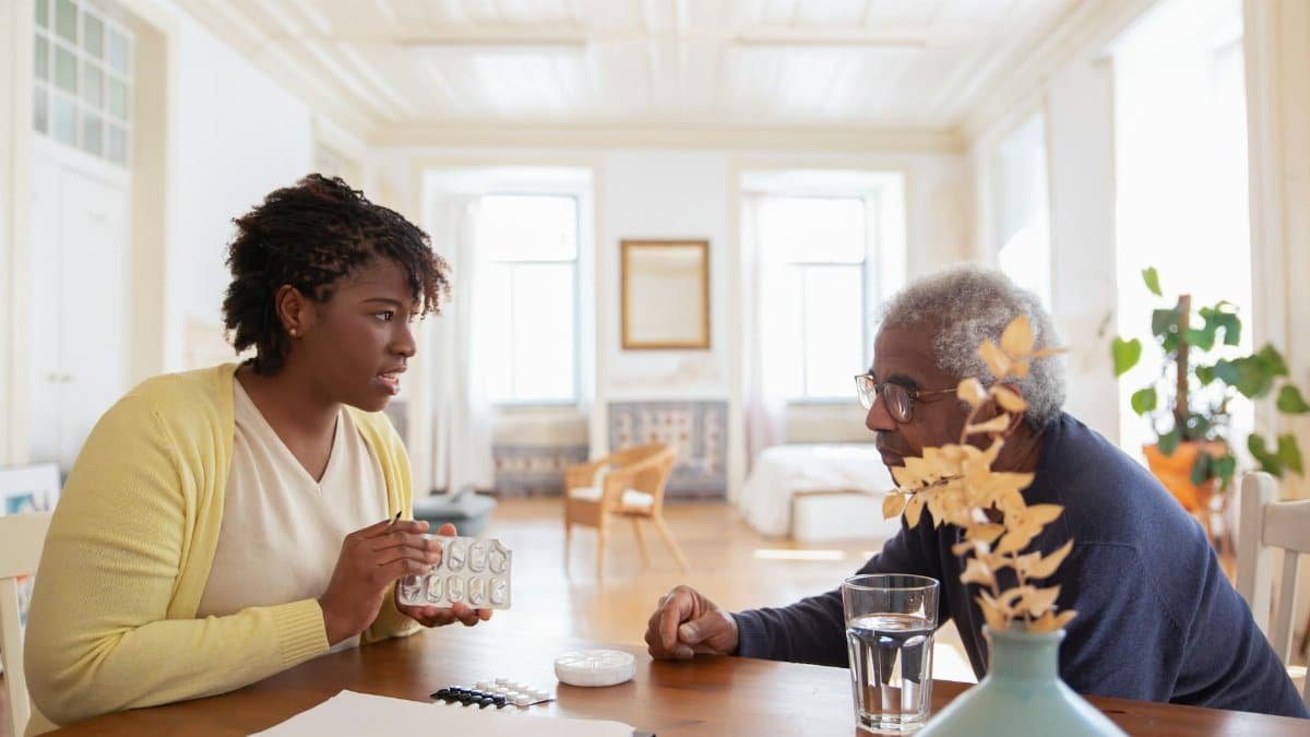 An elderly man sits with a caregiver discussing medication at a table in a bright room.