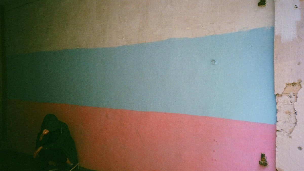 A person in a hooded sweater crouches against a vibrant, textured wall.