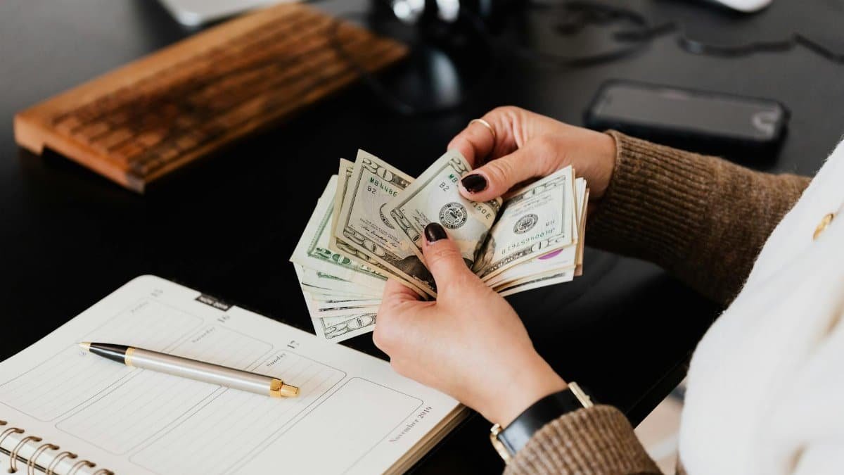 Unrecognizable elegant female in sweater counting dollar bills while sitting at wooden table with planner and pen