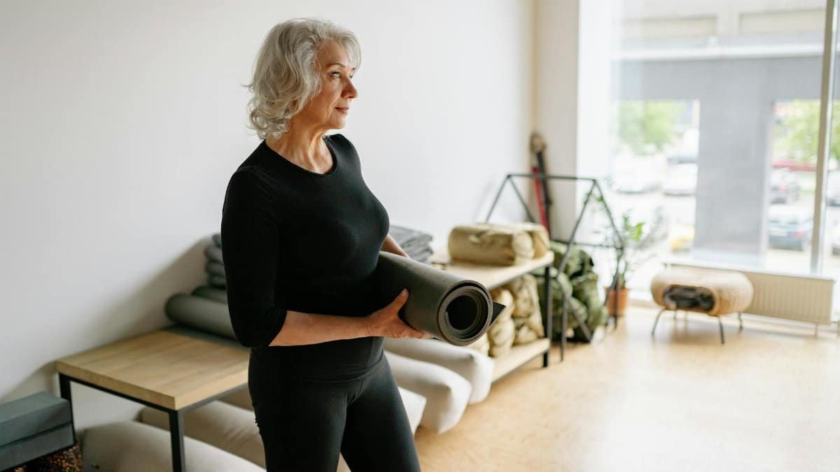 Elderly woman in black attire holding a yoga mat in a bright, minimalist space.