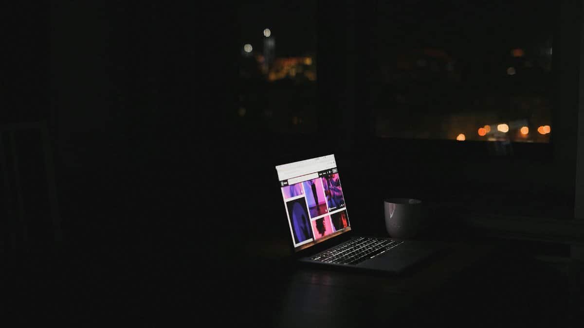 A laptop glows in a dark room at night with a cityscape through the window. Ideal for tech and solitude themes.