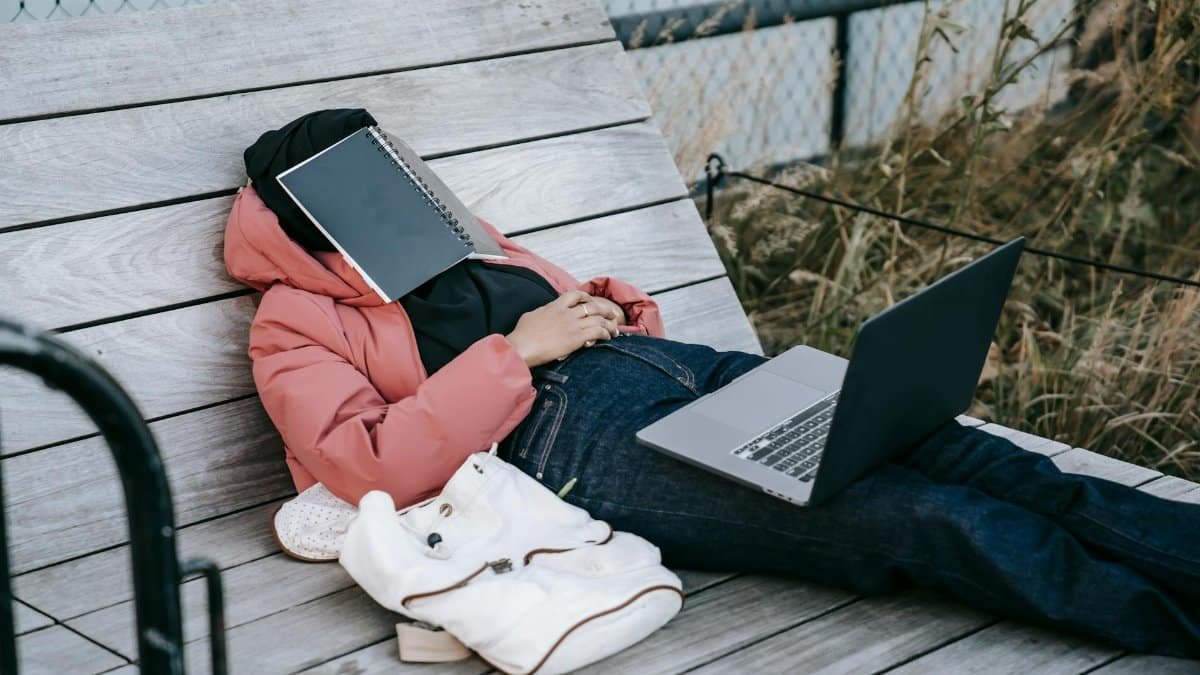 Exhausted student napping on a bench outdoors with a laptop and notepad on face.