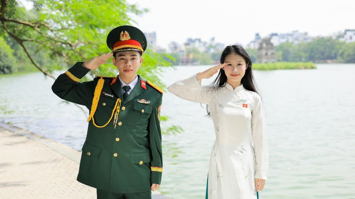 Vietnamese officer and woman in traditional attire saluting by scenic lake in Hanoi.