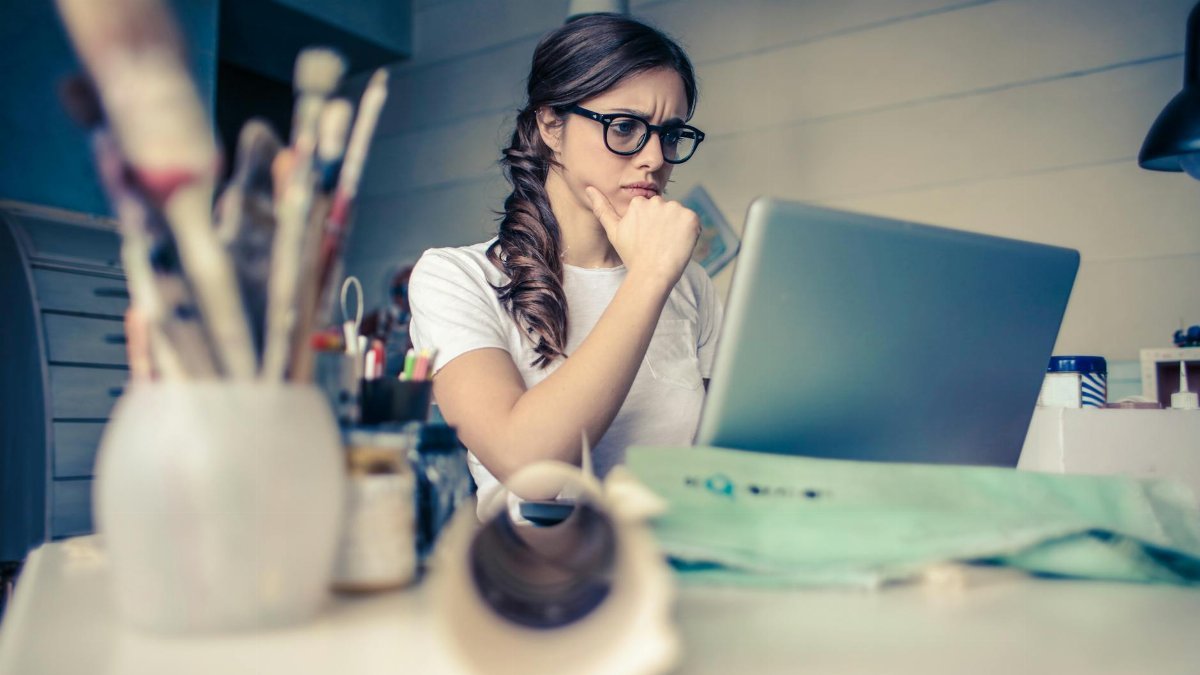 Young woman with glasses deeply focused on a laptop surrounded by art supplies in a home office.