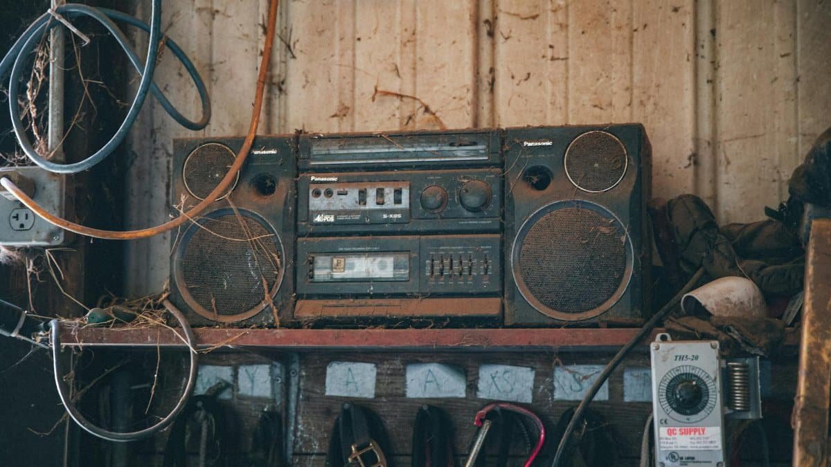 Old fashioned obsolete compact stereo system covered with dust placed on shelf near wooden wall in workshop with tools and wires
