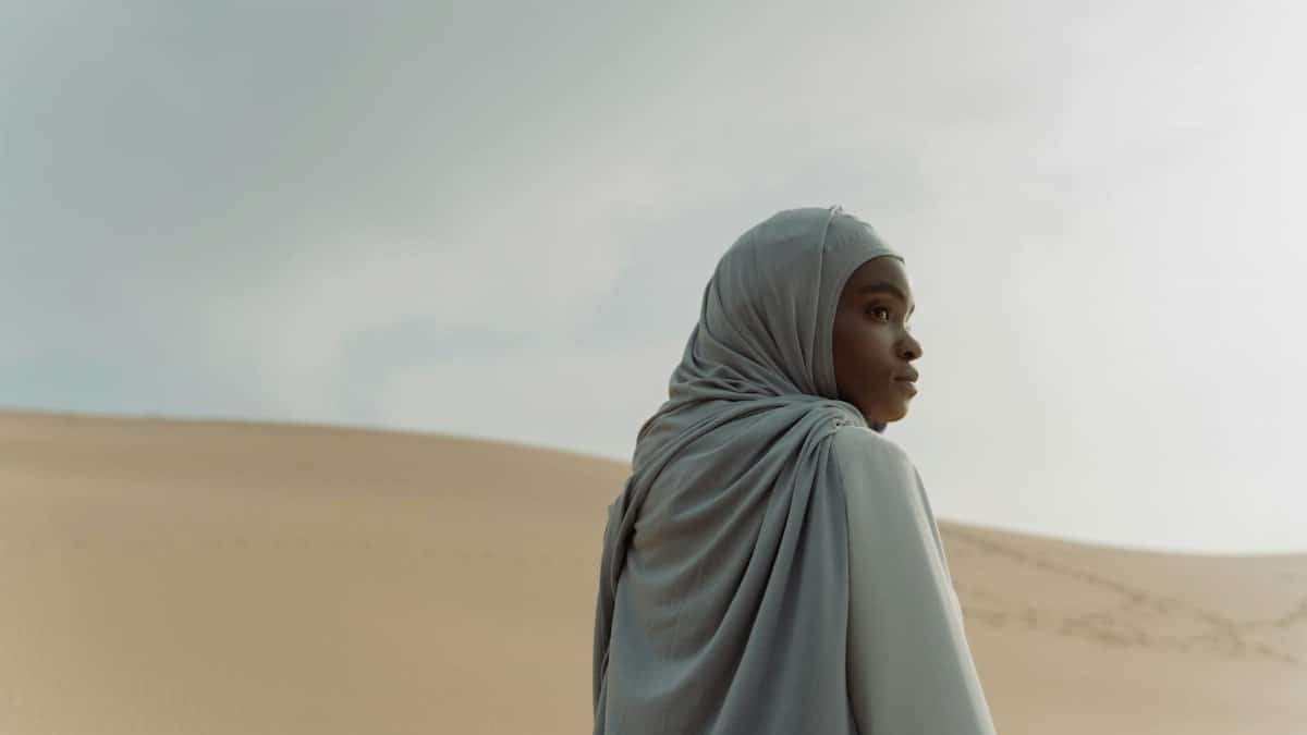 A woman wearing a headscarf stands in the desert, gazing over her shoulder, creating a serene and thoughtful scene.