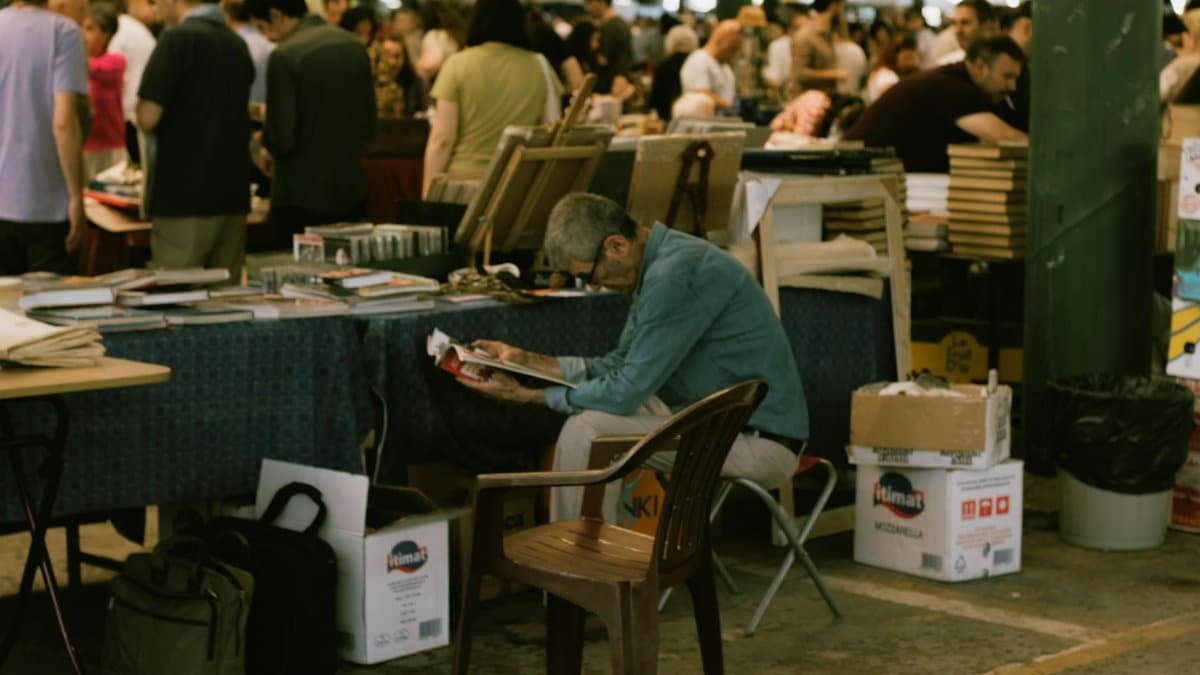 Older man reading a magazine at a bustling outdoor book market.