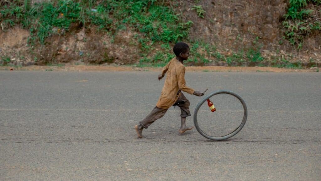 A child joyfully runs alongside a rolling bicycle wheel on a rural road, capturing a moment of simple play.