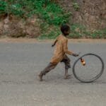 A child joyfully runs alongside a rolling bicycle wheel on a rural road, capturing a moment of simple play.