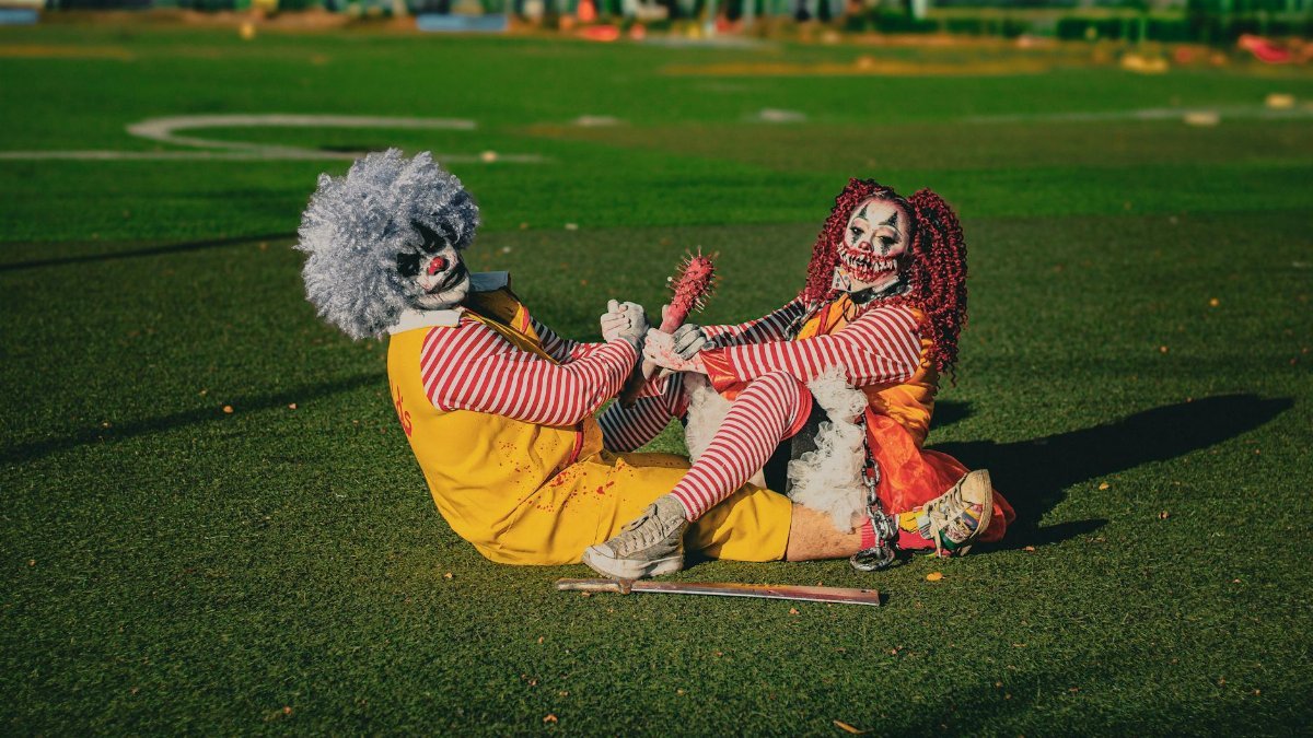 Two frightening clowns dressed in vibrant costumes sitting playfully on a sunny field.