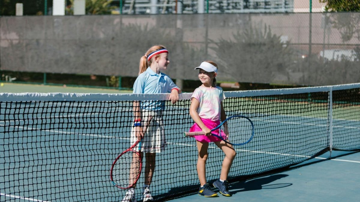 Two young girls in sportswear chatting at the tennis net during a break on an outdoor court.