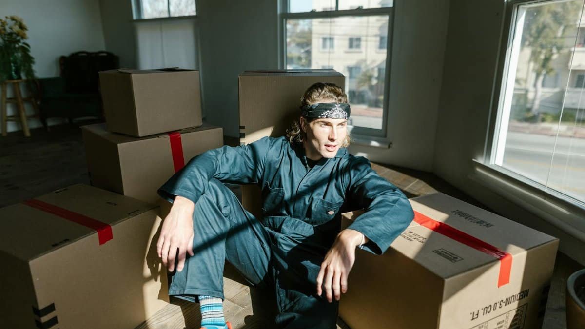 Adult male in jumpsuit sitting on floor amidst cardboard boxes inside a new house.