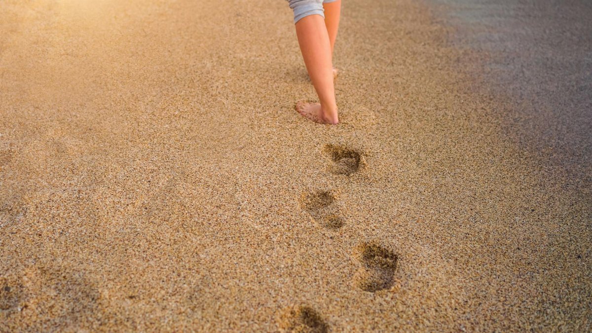 A person walks barefoot on a sandy beach, leaving footprints at sunset.