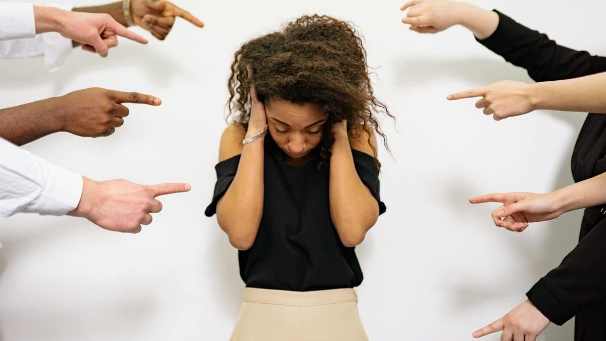 A young woman with curly hair looks distressed as multiple hands point accusatory fingers at her.