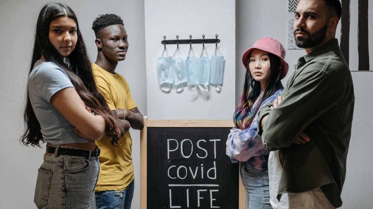 Diverse individuals standing in front of a chalkboard reading 'Post COVID Life' with hanging face masks.