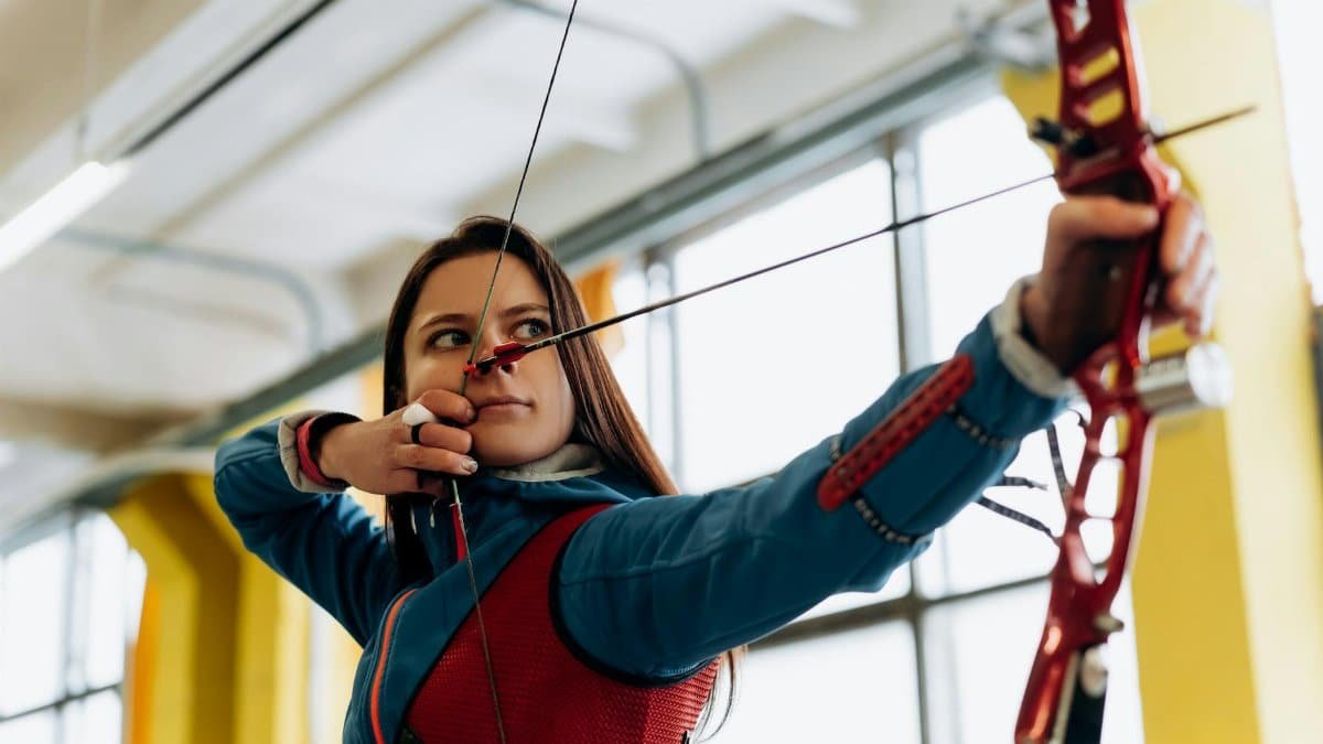 Young female archer in blue aiming a bow and arrow indoors, showcasing concentration and athleticism.