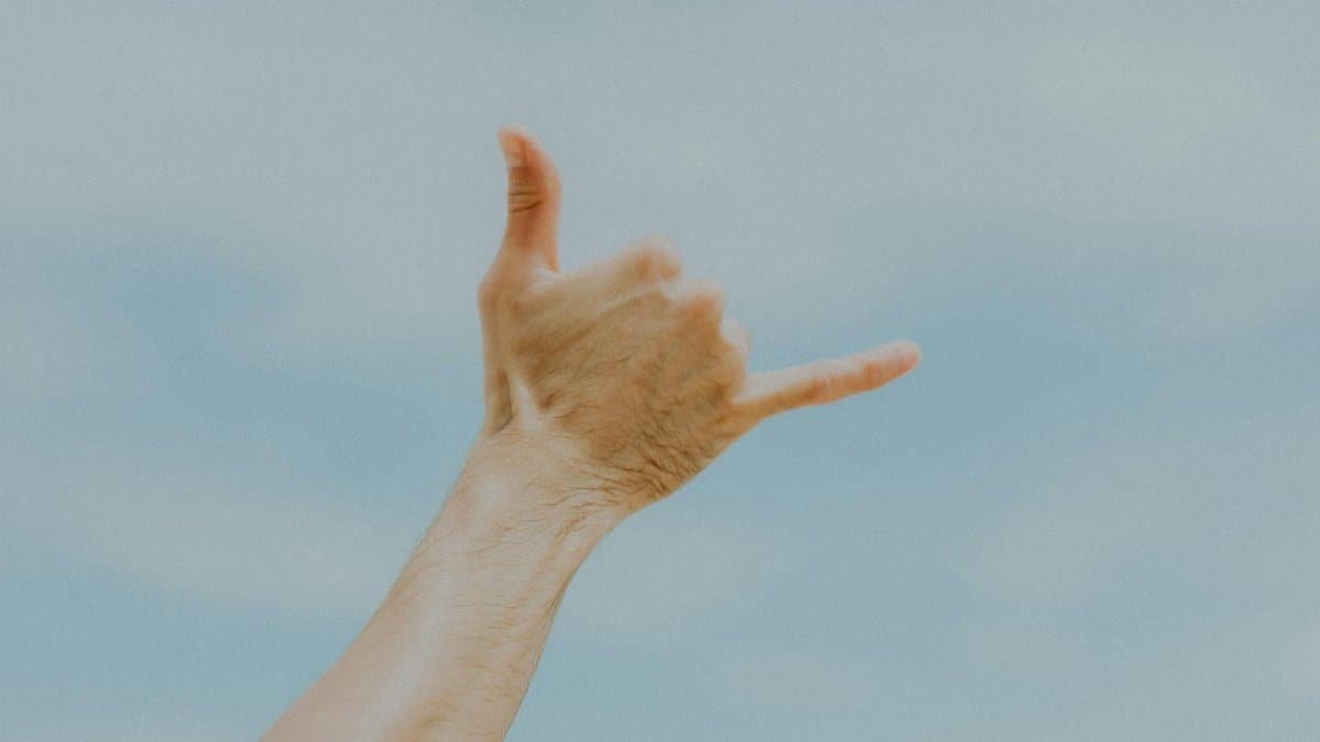 A hand making a shaka gesture in front of a clear blue sky, symbolizing aloha spirit.