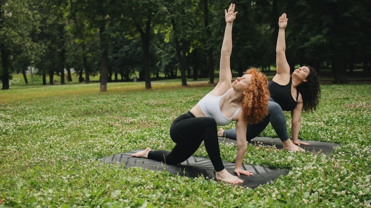 Two women practicing yoga poses on mats in a green park, focused on wellness and mindfulness.