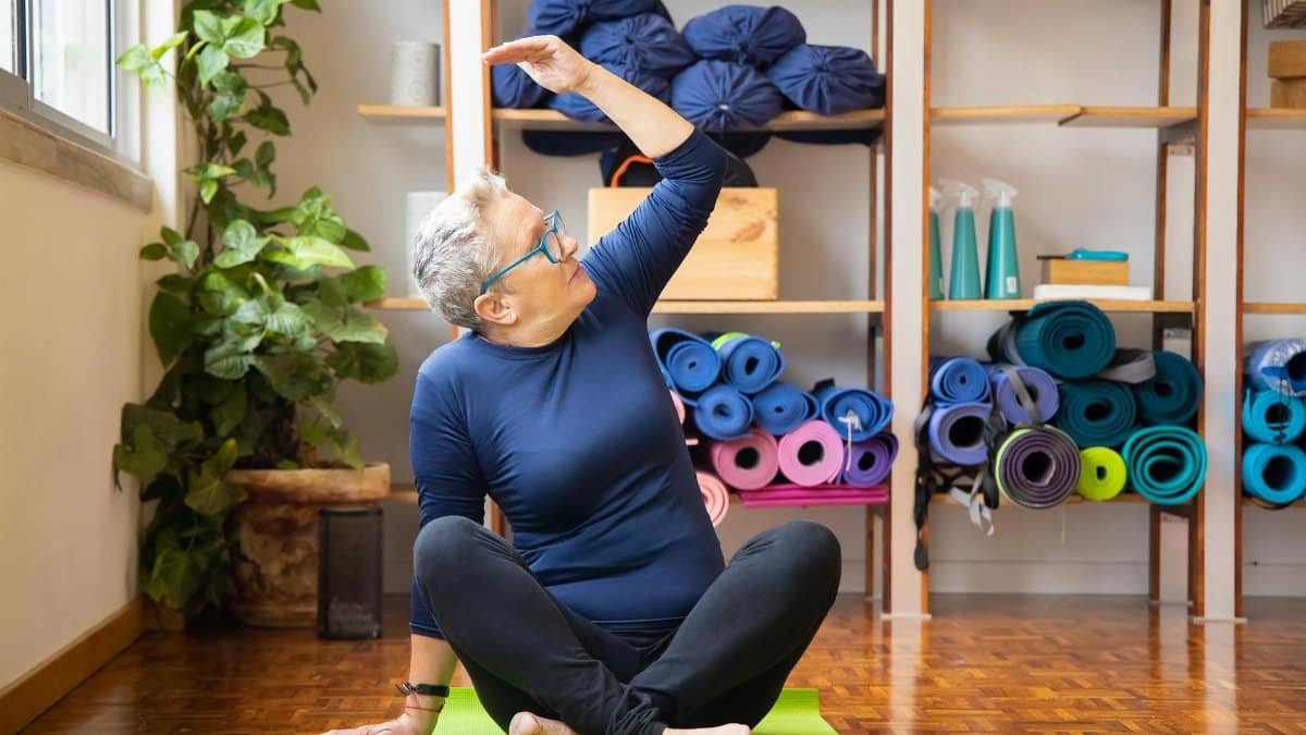 Elderly woman practicing yoga seated indoors on a green mat, promoting healthy lifestyle.