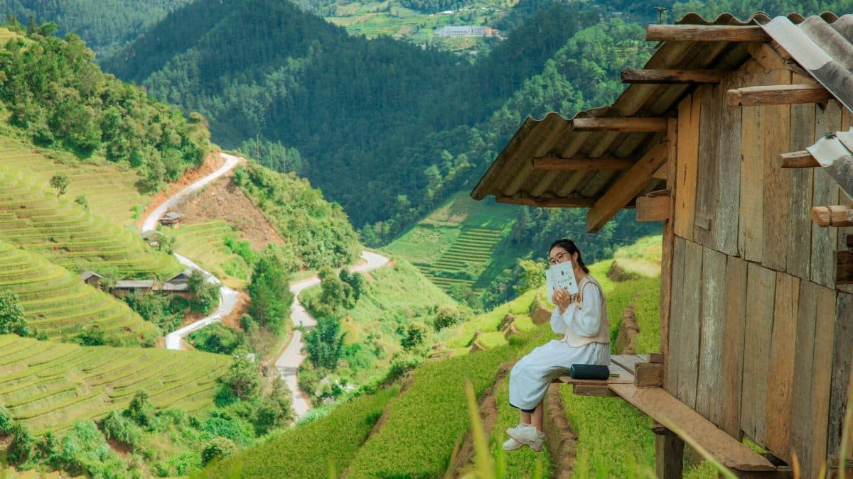 Young woman reads by a rustic hut amidst lush green terraced fields under clear skies.
