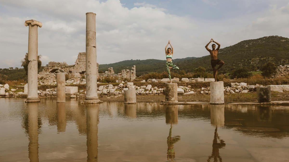 Two people practicing yoga poses on ancient stone ruins beside water. Reflective and serene atmosphere.