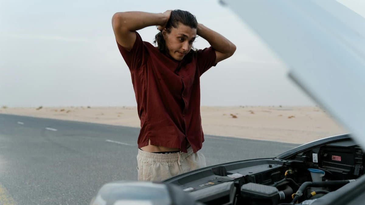 A frustrated man examining a broken car engine on a deserted road, showing stress and concern.