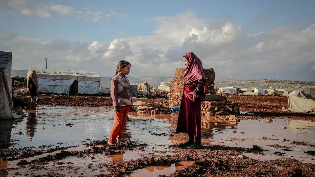 Two children talk beside puddles in an Idlib refugee camp, surrounded by tents and muddy terrain.
