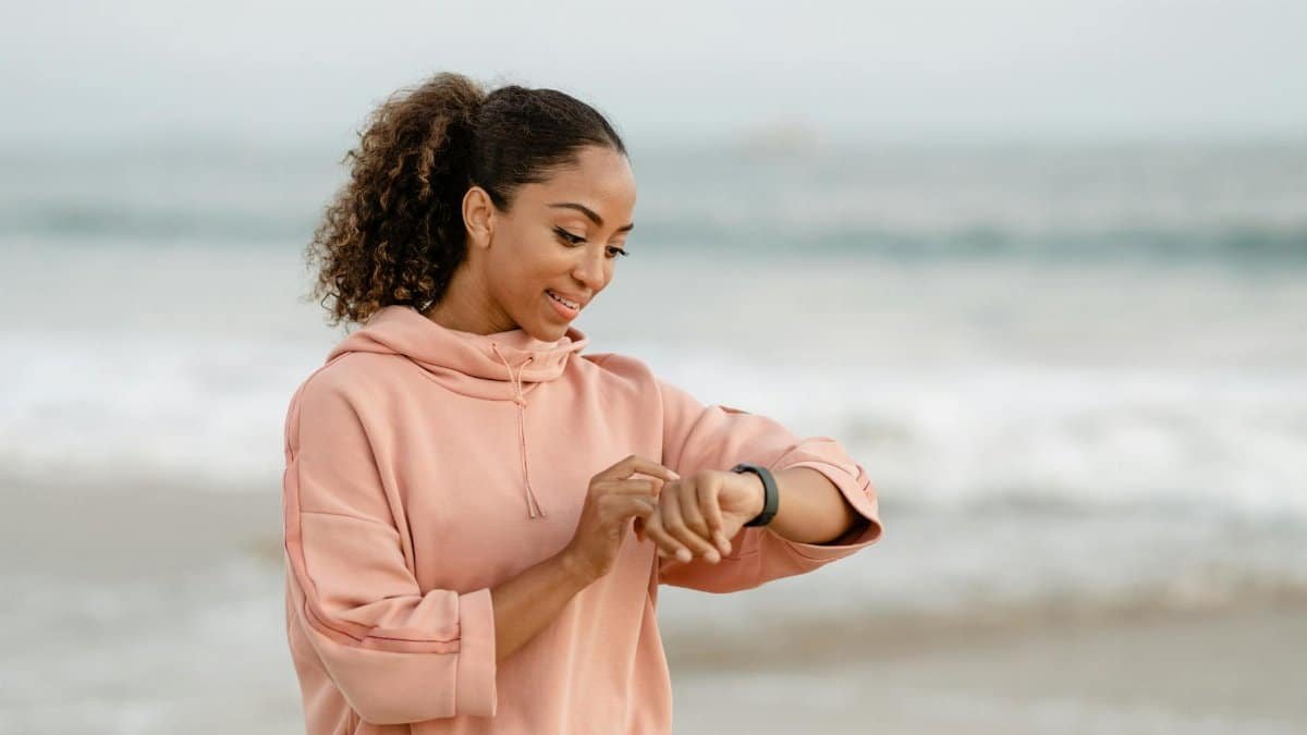 Smiling woman in a pink hoodie checks her smartwatch by the ocean.