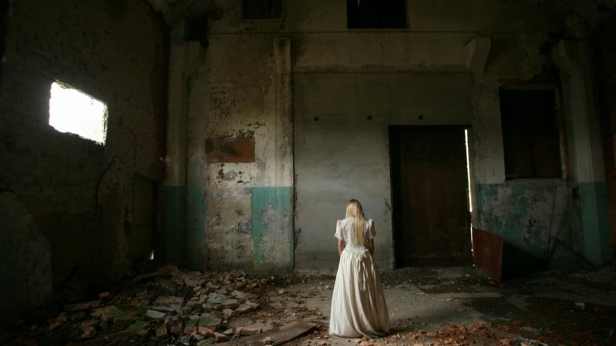 A woman in white stands in an eerie, abandoned building with decayed walls.