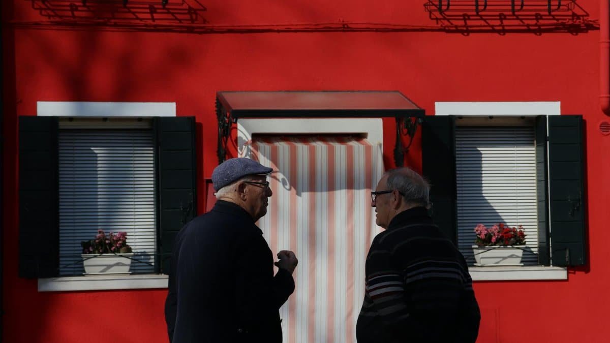 Two senior men talking in front of a bright red building with white accents and flower boxes.