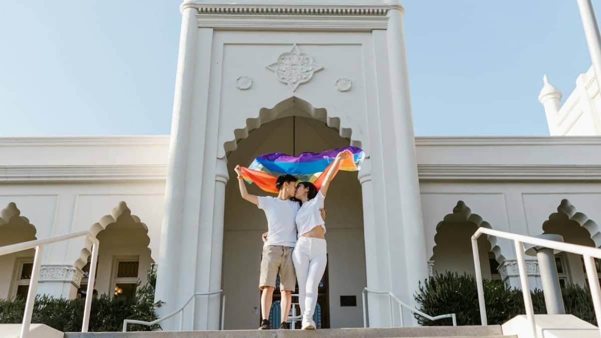 A loving LGBTQ+ couple celebrates happiness holding a rainbow flag in front of an iconic building.
