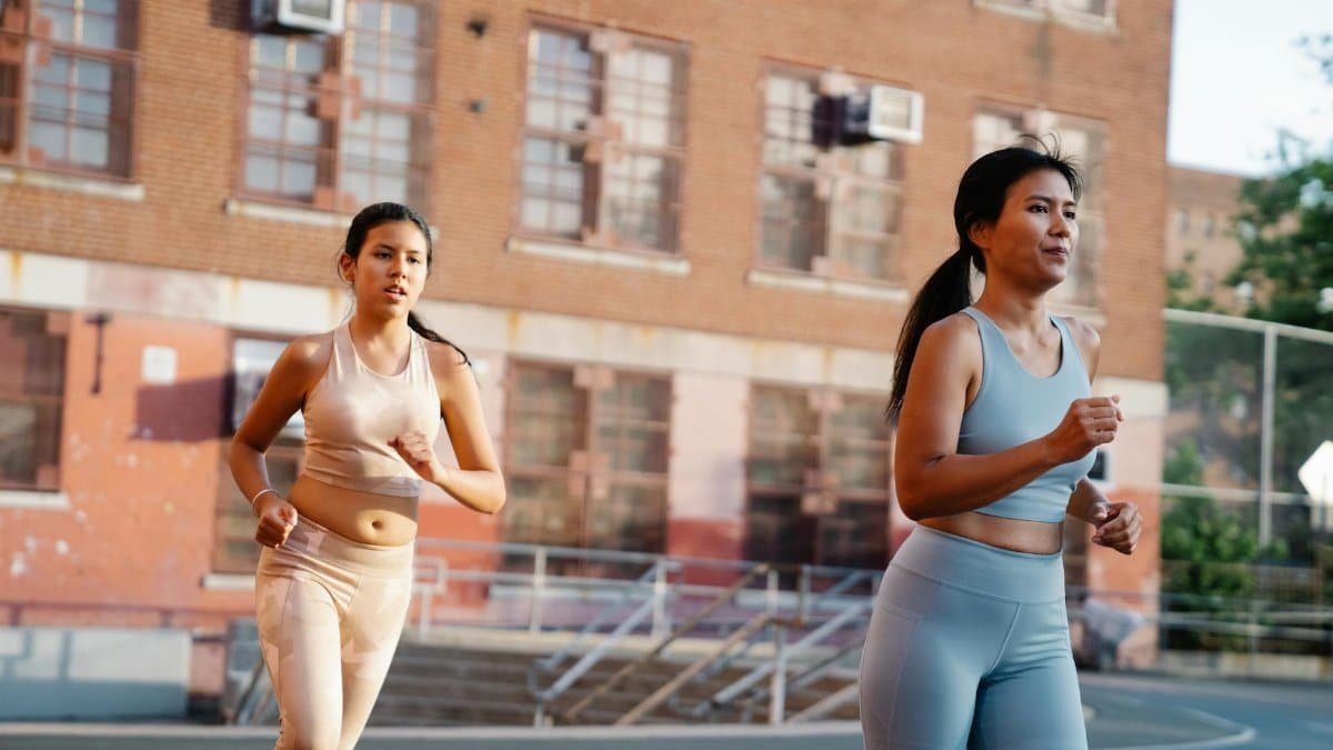 Two women jogging on an outdoor court near a brick building in activewear.
