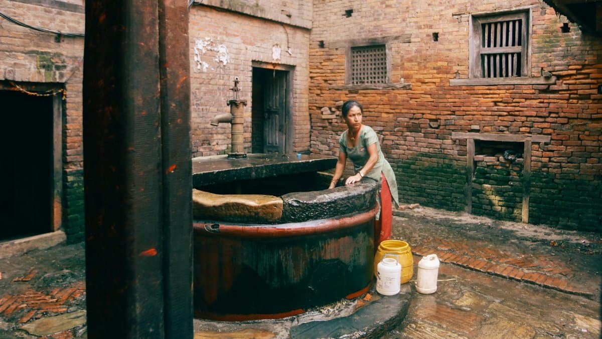 A woman draws water from a traditional well in a residential courtyard with brick walls.