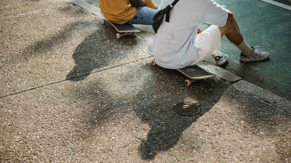 From above of crop anonymous multiethnic male friends sitting on skateboard while spending free time on asphalt road on summer sunny day