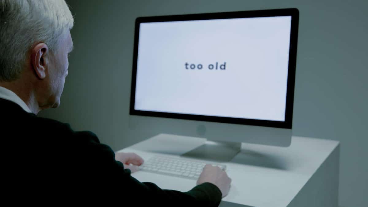 Senior man at computer facing ageism online, "too old" displayed on screen.
