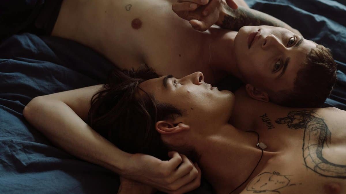 Two tattooed men embracing intimately in a bedroom setting.