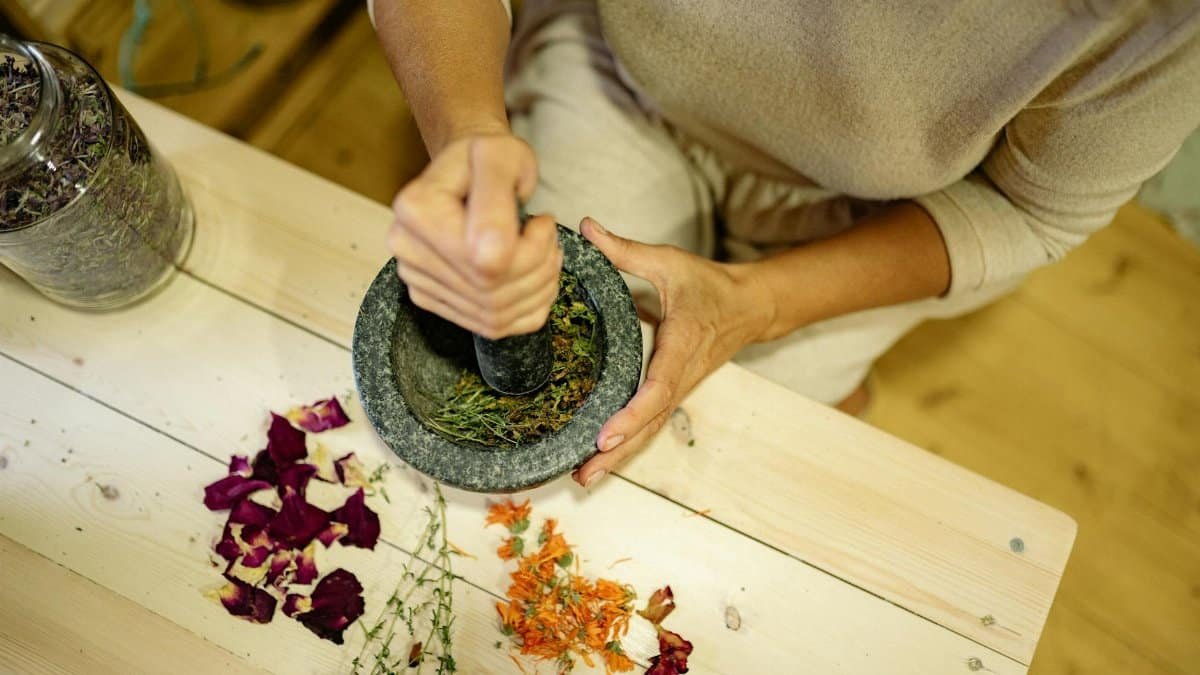 Close-up of hands grinding herbs and flowers in a mortar on a wooden table.