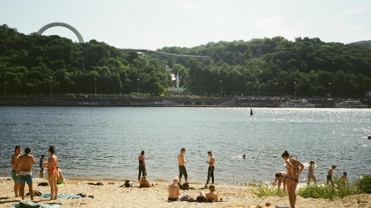 People enjoying a sunny day on a sandy beach by the river in Kyiv, Ukraine, with iconic landmarks in the background.