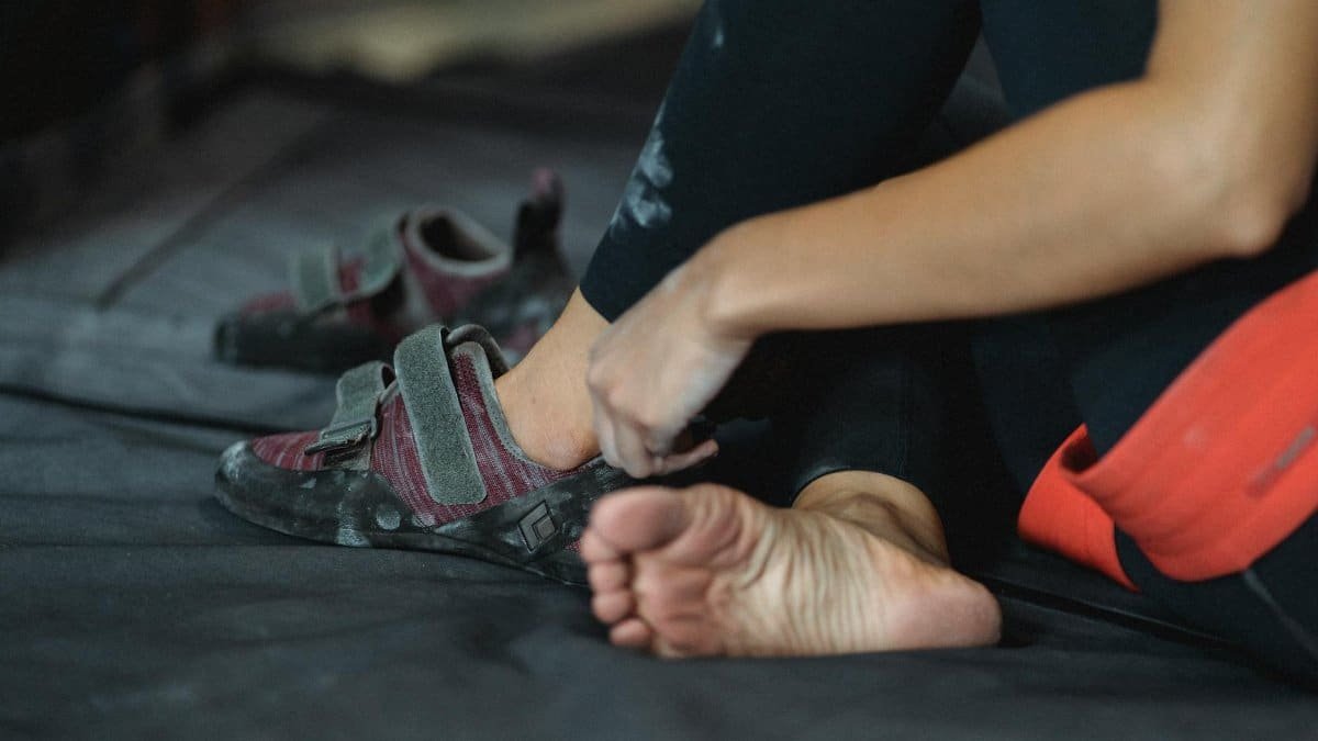 Side view of unrecognizable barefoot mountaineer sitting on mat with crossed legs while putting on rock climbing shoes during training in gym on blurred background