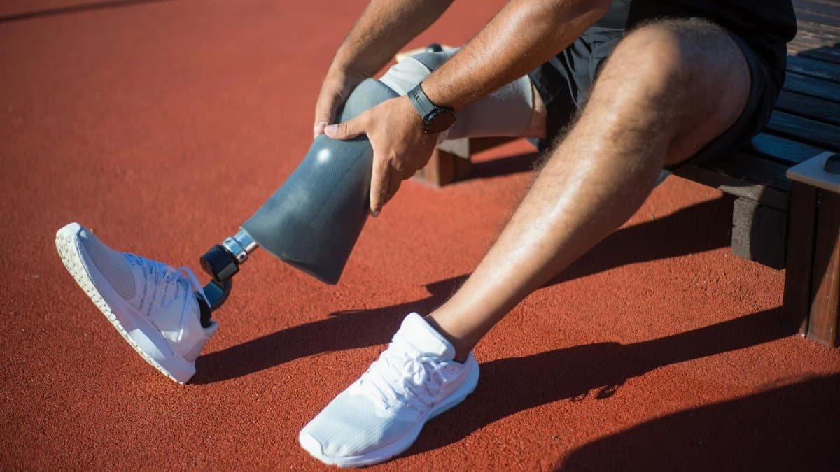 An adult man sitting on a bench adjusting his prosthetic leg on an outdoor track.