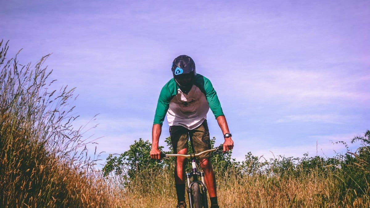 A mountain biker rides through a grassy trail in Nelson, New Zealand, wearing a helmet and casual attire.