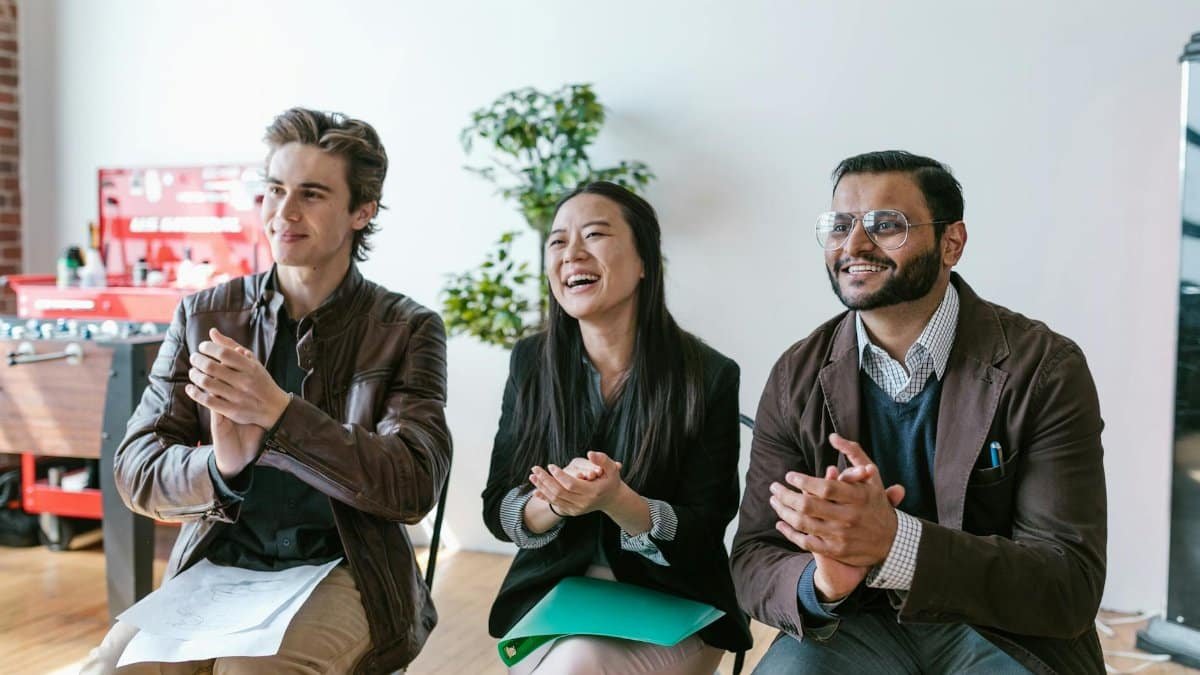 Three diverse team members clapping and smiling during an indoor meeting, showing positivity and teamwork.