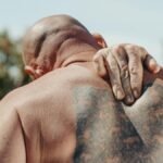 Bald man with tattoo holding back in pain under sunny conditions.