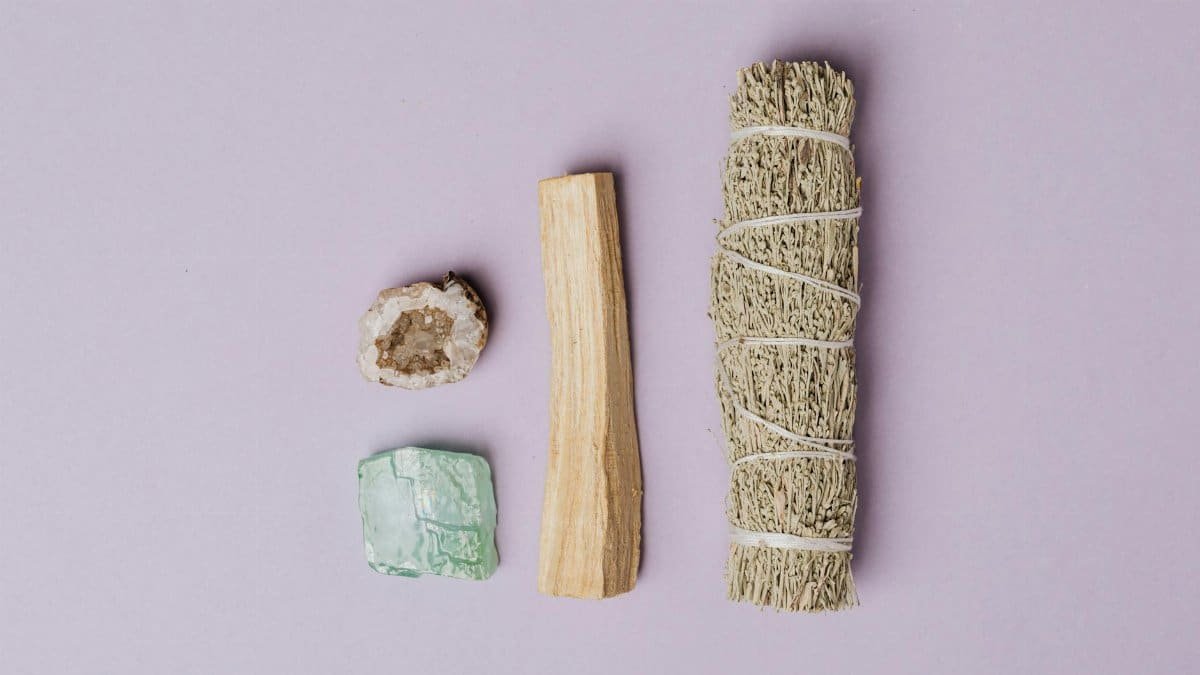 Flat lay of spiritual healing tools including sage, palo santo, quartz geode, and amazonite on a lavender background.
