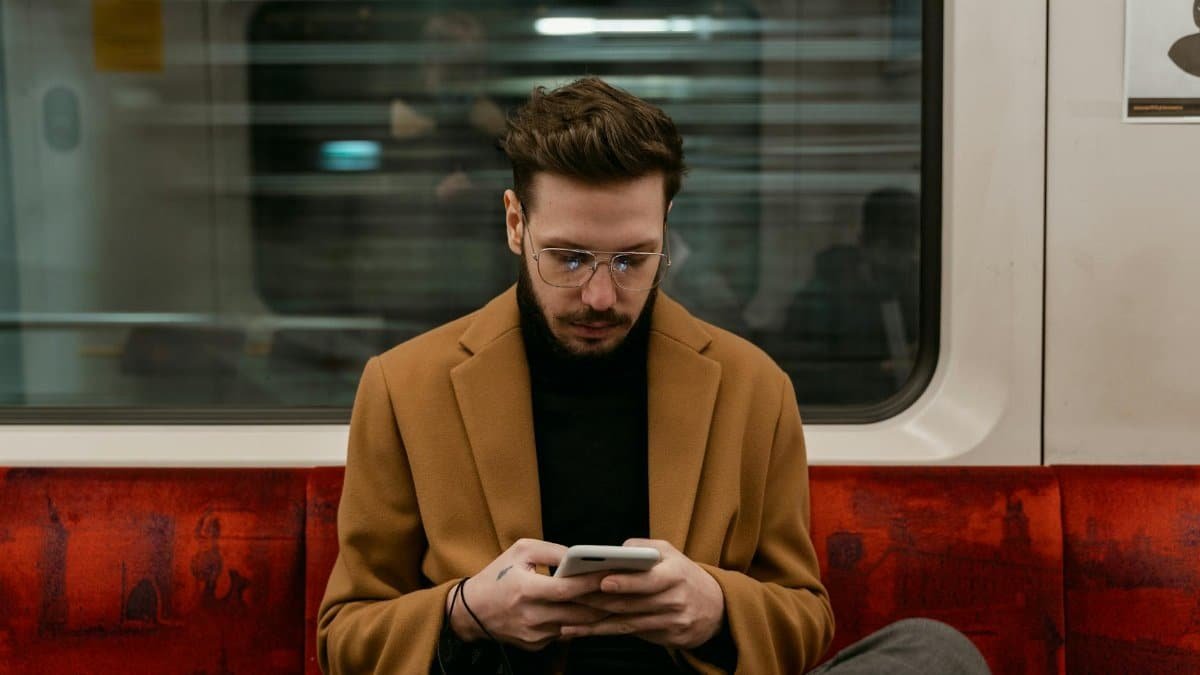 A man in a brown coat uses his smartphone while commuting on a train. Urban city life captured indoors.