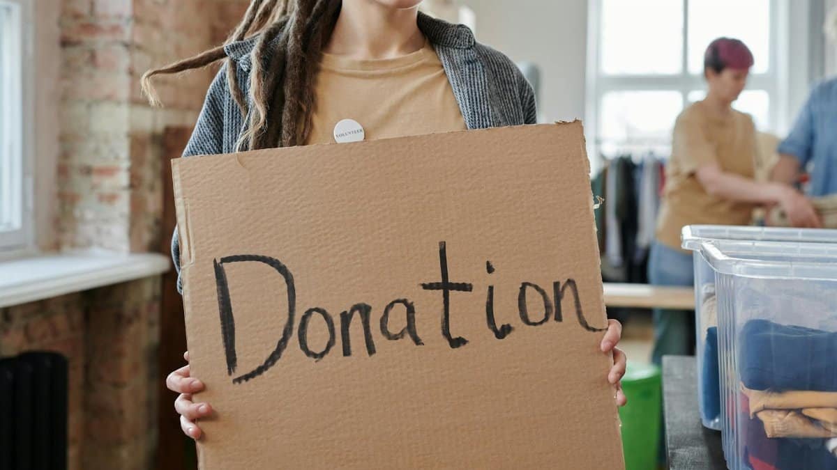 Volunteer holding a cardboard donation sign in a community center with clothing and supplies.