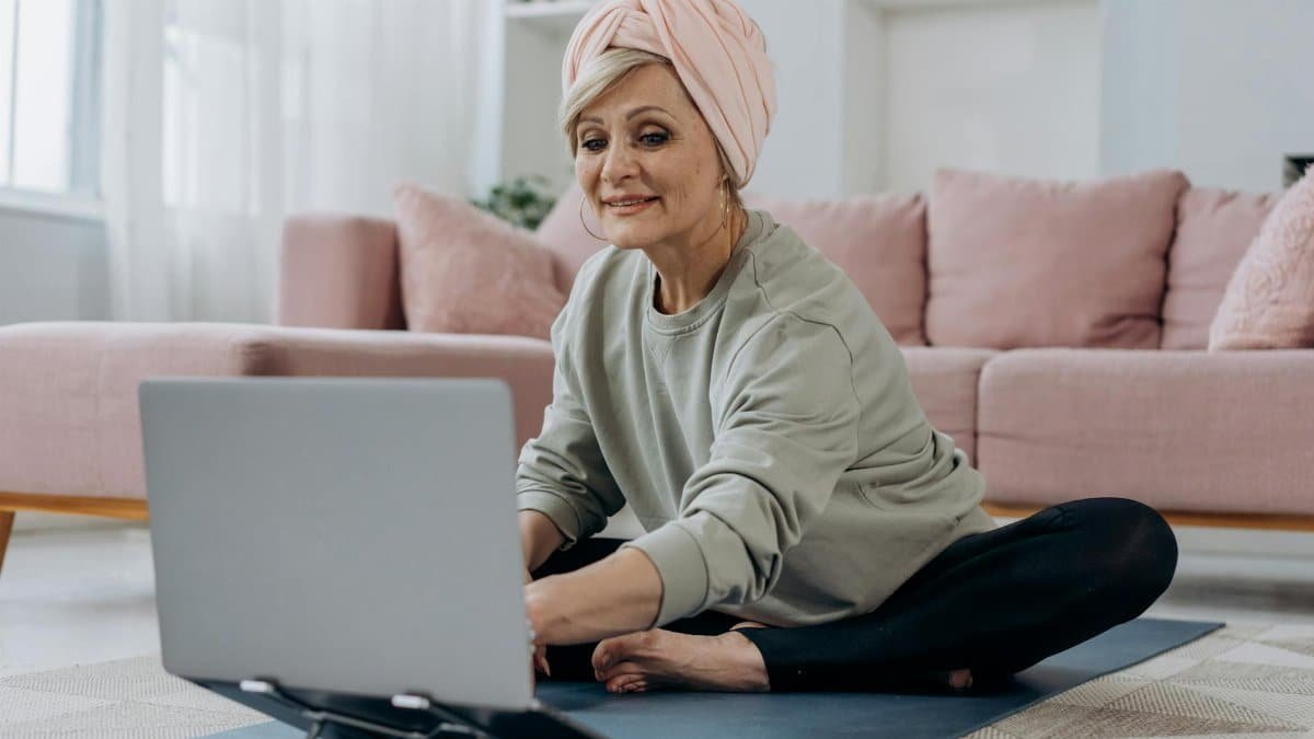 Senior woman using a laptop for an online yoga class, sitting indoors on a yoga mat with a relaxed expression.