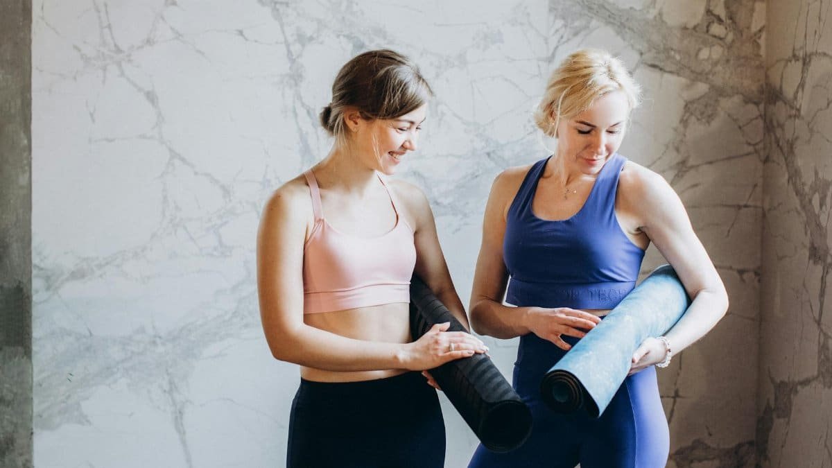 Two women in activewear holding yoga mats, smiling and preparing for a fitness session.