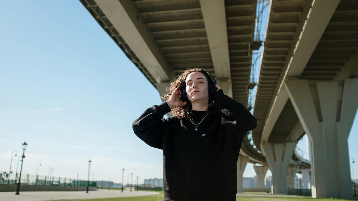 Youthful woman listens to music under a modern bridge, experiencing solitude in a cityscape.